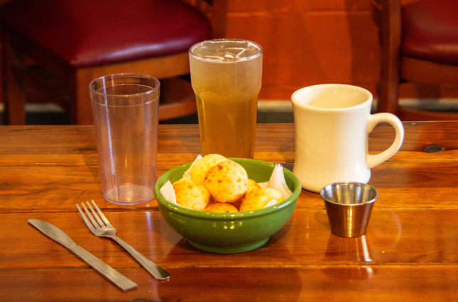 Ceramic bowl with pao de queijo surrounded by reusable cups, silverware, and sauce cups