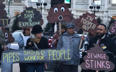 A crowd of people rallying outside of Baltimore City Hall, with signs like "Pipes should work for people" and "sewage stinks"