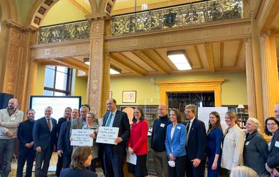 Elizabeth Saunders (in red) stands with Senator Jamie Eldridge, Representative Ruth Balser (holding signs), and members of the Public Lands Preservation Act coalition at a celebration of the signing of the bill in Januar