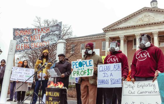 People rallying in front of the MD State House, some wearing gas masks, with signs like "Burning trash is not clean energy," "clean air is a human right," and "people over profit."