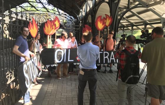 15 people hold signs saying "Stop Oil Trains" and showing train tank cars and flames as news media films